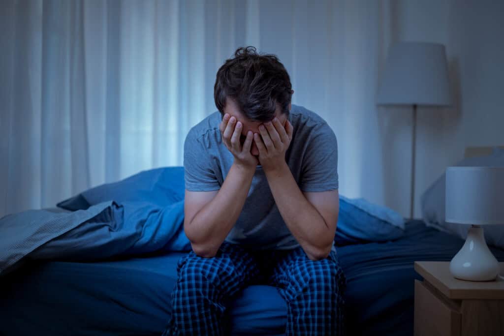 Man sitting on bed with head in hands, appearing distressed or anxious.