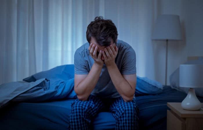 Man sitting on bed with head in hands, appearing distressed or anxious.