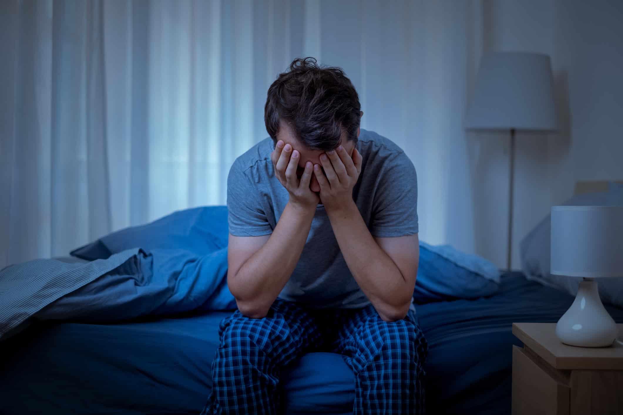 Man sitting on bed with head in hands, appearing distressed or anxious.