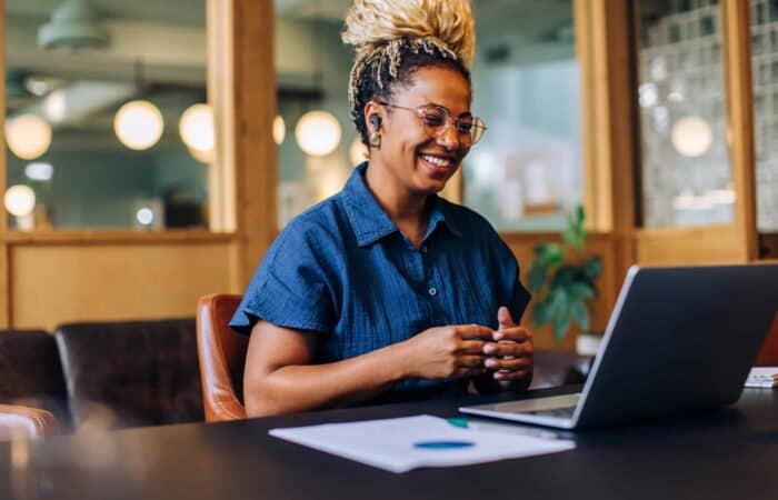 Smiling woman with glasses working on a laptop in a modern office setting.